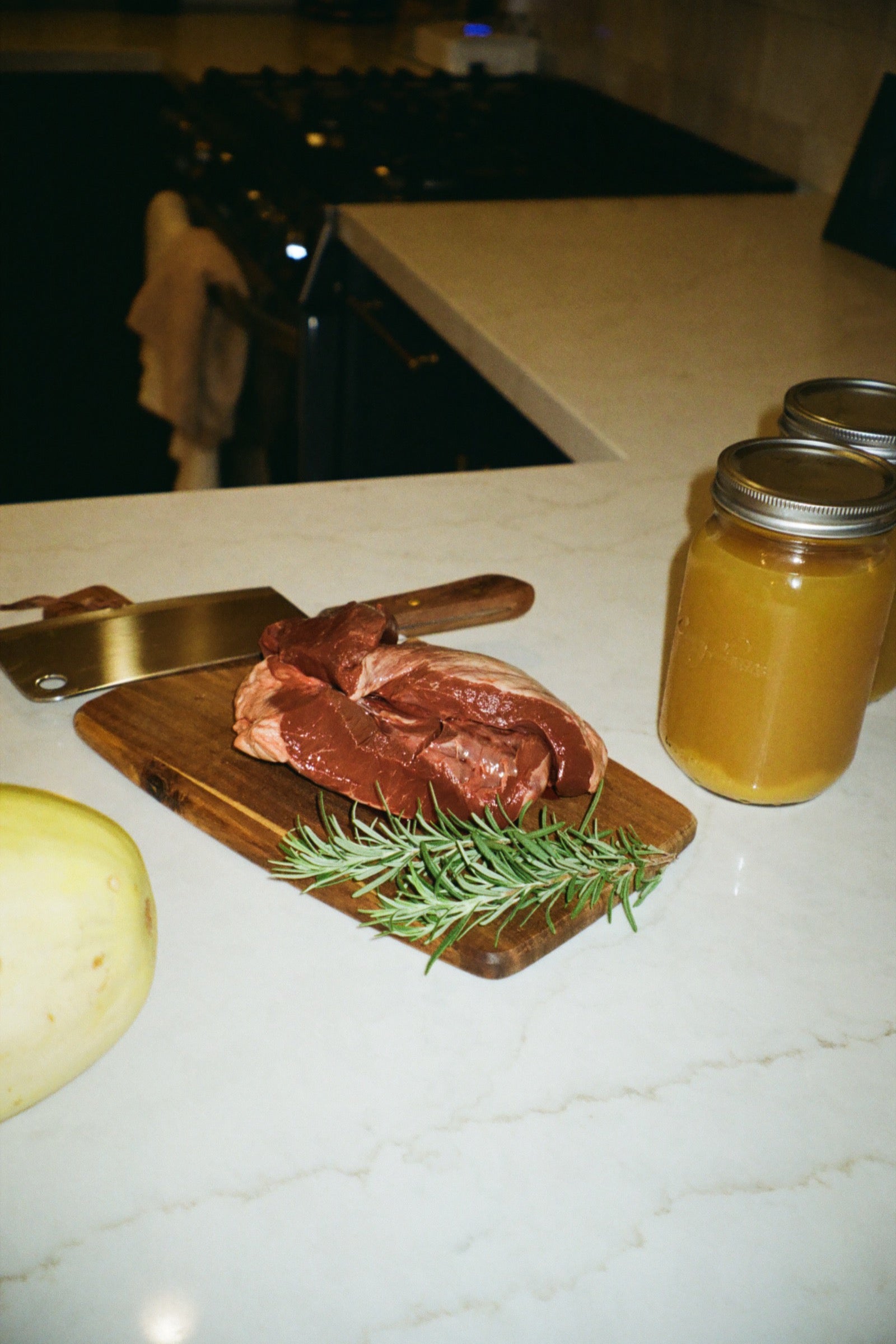 Raw meat, a cleaver, rosemary, and jars arranged on a kitchen counter